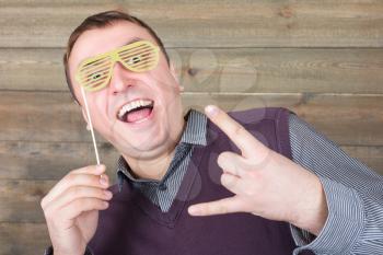 Playful young man with funny hipster sunglasses on a stick, wooden background. Fun photo props and accessories for shoots