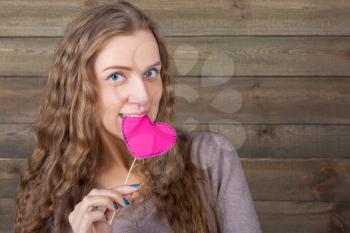 Cute woman with funny heart on a stick, wooden background. Fun photo props and accessories for photo shoots