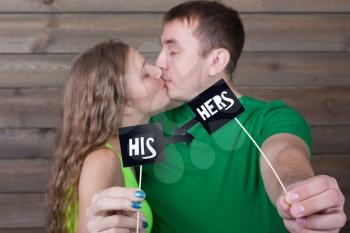 Love couple kisses and shows funny icons on a sticks with just married inscription, wooden background. Fun photo props and accessories for shoots