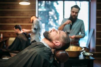 Bearded man in salon cape at the barbershop, barber on background