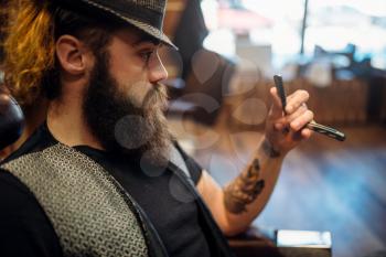 Bearded barber in hat with straight razor in hand, barbershop inerior on background