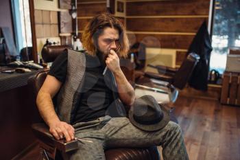 Thoughtful bearded barber sitting in a leather chair, barbershop inerior on background