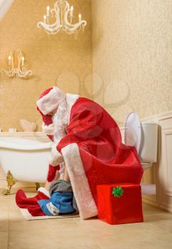 Santa Claus with his pants down sitting on the toilet, gift box in wrapping paper on background. Christmas humor