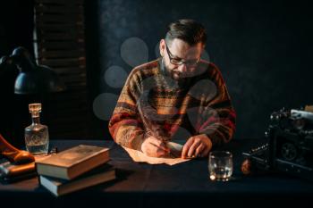 Bearded writer in glasses writes with a feather. Retro typewriter, crystal decanter, books and vintage lamp on the desk