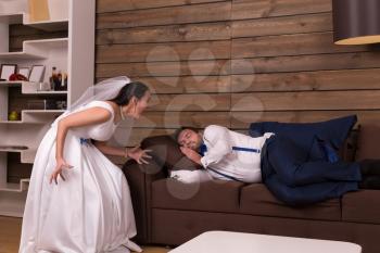 Bride in white dress and veil is shouting on a drunk sleeping on couch groom. After wedding celebration. Wooden interior of the room on background