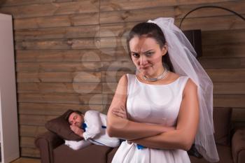 Unhappy bride in white dress and veil, sleeping on couch groom on background. After wedding celebration