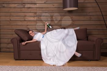 Drunk bride with bottle of alcohol in hand relax on couch after wedding celebration. Wooden interior of the room on background
