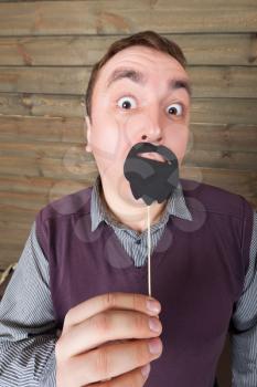 Playful male with funny sunglasses and beard on a stick, wooden background. Fun photo props and accessories for shoots