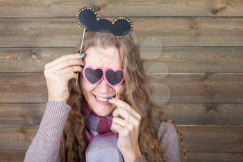 Cute girl with funny hairstyle and sunglasses in form of hearts on a sticks, wooden background. Fun photo props and accessories for shoots
