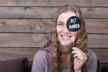 Cute smiling girl shows funny icon on a stick with just married inscription, wooden background. Fun photo props and accessories for photo shoots