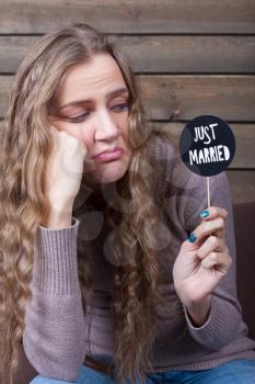 Young woman with sad face holding funny icon on a stick with just married inscription, wooden background. Fun photo props and accessories for shoots