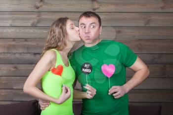 Woman kisses man and shows funny icons on a sticks with hearts and just married inscription, wooden background. Fun photo props and accessories for shoots