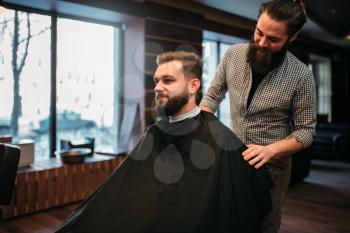 Bearded man in salon cape at the barbershop, barber prepares to cut