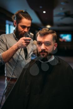 Barber working by clipper with hairstyle of the client man in black salon cape, barbershop on background