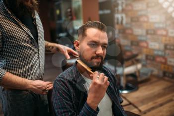 Male client combing his beard at the barbershop, professional barber on background