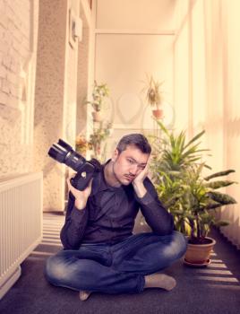Thoughtful photographer with camera sitting in a yoga pose, house plants on background