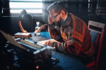 Bearded journalist in glasses reading his work. Retro typewriter, feather, crystal decanter, books and vintage lamp on the desk
