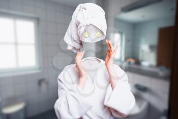 Girl in white bathrobe with towel on head with cucumbers on eyes touch her invisible face. Invisibility fantasy concept, transparent female person. Personal hygiene advertising or marketing