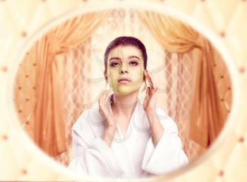 Young woman in white bathrobe gets a cream mask for the face against mirror in bedroom. Girl take care of beaty