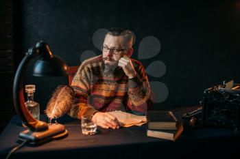 Thoughtful bearded writer in glasses sitting at the table. Retro typewriter, feather, crystal decanter, books and vintage lamp on the desk