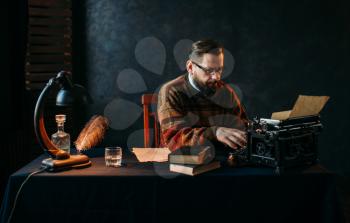 Thoughtful bearded writer in glasses typing on vintage typewriter. Retro feather, crystal decanter, books and lamp on the desk