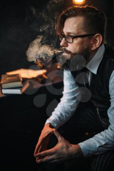 Portrait of serious bearded man in glasses smoking pipe, table with books on background. Writer, journalist, literature author, blogger or poet concept