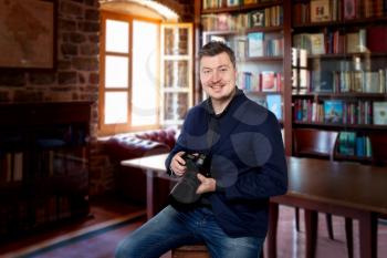 Smiling photographer with digital camera sitting on a chair, home library on background