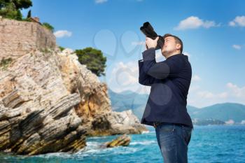 Cameraman in a jacket with digital camera taking picture, rocky coast on background