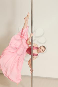 Young sexual poledance woman stretching in beautiful pink dress. Professional strip dancer exercising with pole in dance studio