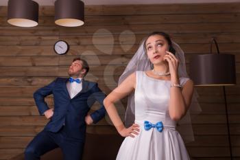 Portrait of bride using mobile phone and groom posing on camera, wooden room on background.