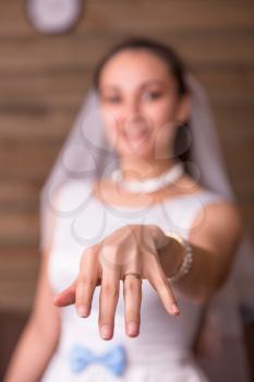 Bride in white dress and veil showing golden wedding ring on her hand, wooden room interior on background