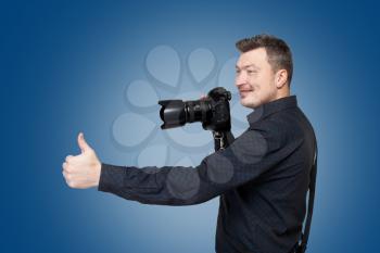 Photographer with professional digital camera shows thumb up, blue background