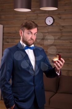Success groom in luxury suit holding box with wedding ring in his hand, wooden room interior on background
