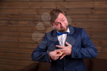 Groom in suit and bow-tie trying to put on a wedding ring on his finger, wooden room interior on background