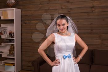 Cute bride in veil and white dress with blue bow posing, wooden room interior on background