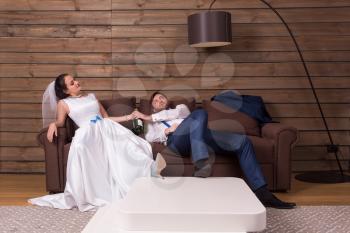 Drunk bride and groom relax on couch with bottle of alcohol after wedding celebration. Wooden interior of the room on background