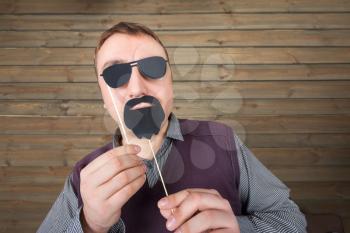 Playful male with funny sunglasses and beard on a stick, wooden background. Fun photo props and accessories for shoots