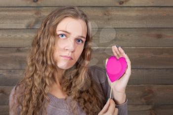 Young female with funny pink heart on a stick, wooden background. Fun photo props and accessories for shoots