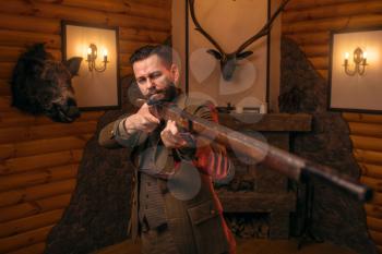 Hunter man aims of the antique hunting rifle. Fireplace, stuffed wild animals, bear skin and other trophies on background