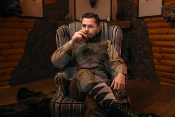 Hunter man in traditional vintage hunting clothing drink luxury alcohol after successful hunt. Fireplace, stuffed wild animals, bear skin and other trophies on background