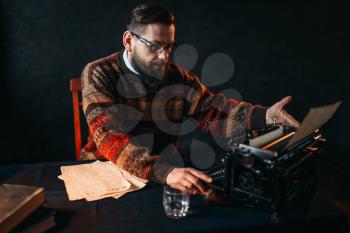 Bearded poet in glasses typing on retro typewriter. Books on the desk with vintage dark blue table cloth