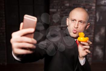 Special agent makes selfie with little toy duck on phone camera. Contract killer in suit and red tie shows his fears and secrets. Hired murderer wallpaper or background concept