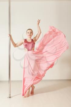 Sexy slim strip dancer exercising with pole in dance studio. Attractive professional poledance girl posing in beautiful pink dress.