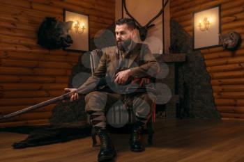 Hunter man in vintage hunting clothing sitting in a chair with antique rifle. Fireplace, stuffed wild animals, bear skin and other trophies on background. Hunt lifestyle