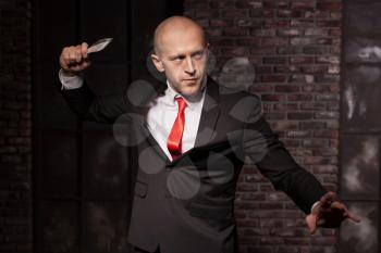 Silent assassin, oriental martial arts in action. Bald contract murderer in suit and red tie holds combat knife. Fighting knife is a dangerous weapon in right hands