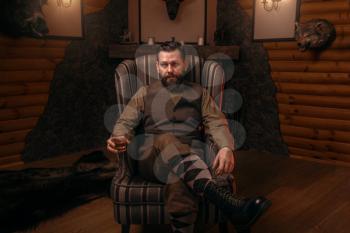 Hunter man in traditional british clothing sitting in a chair after hunting and drink whiskey. Fireplace, stuffed wild animals, bear skin and other trophies on background. Hunt lifestyle