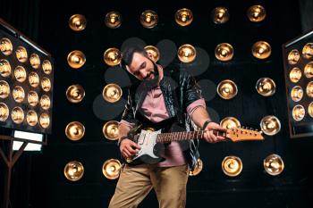 Male bearded guitarist in black leather jacket on the stage with the decorations of lights. Performer with electro guitar, solo concert