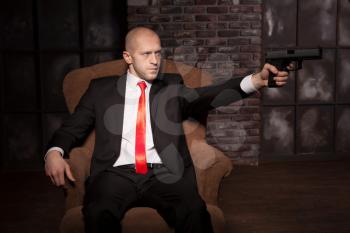 Bald killer in suit and red tie sitting in chair and aims a pistol. Professional secret agent concept