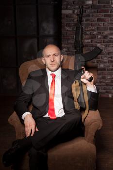 Contract killer in suit and red tie sitting in a chair and holds automatic weapon. Brave professional secret agent on mission