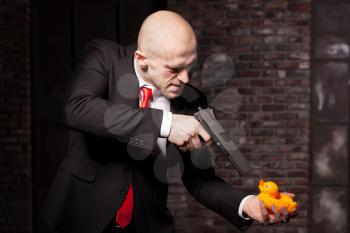 Angry contract murderer in suit and red tie aims a pistol on little toy duck. Professional secret agent concept